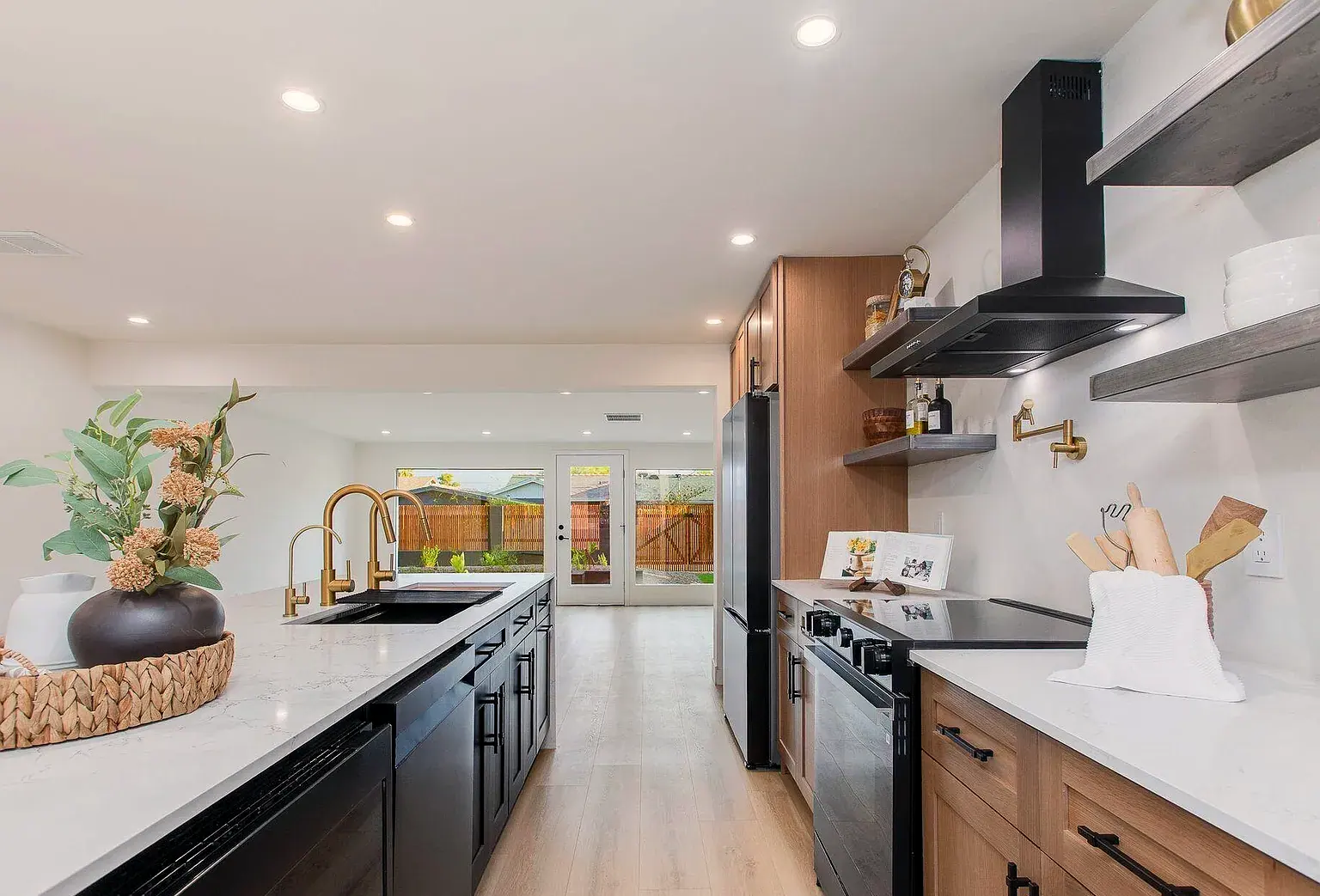 Galley kitchen running toward the yard: black island cabinetry, brass gooseneck faucets, oak uppers.