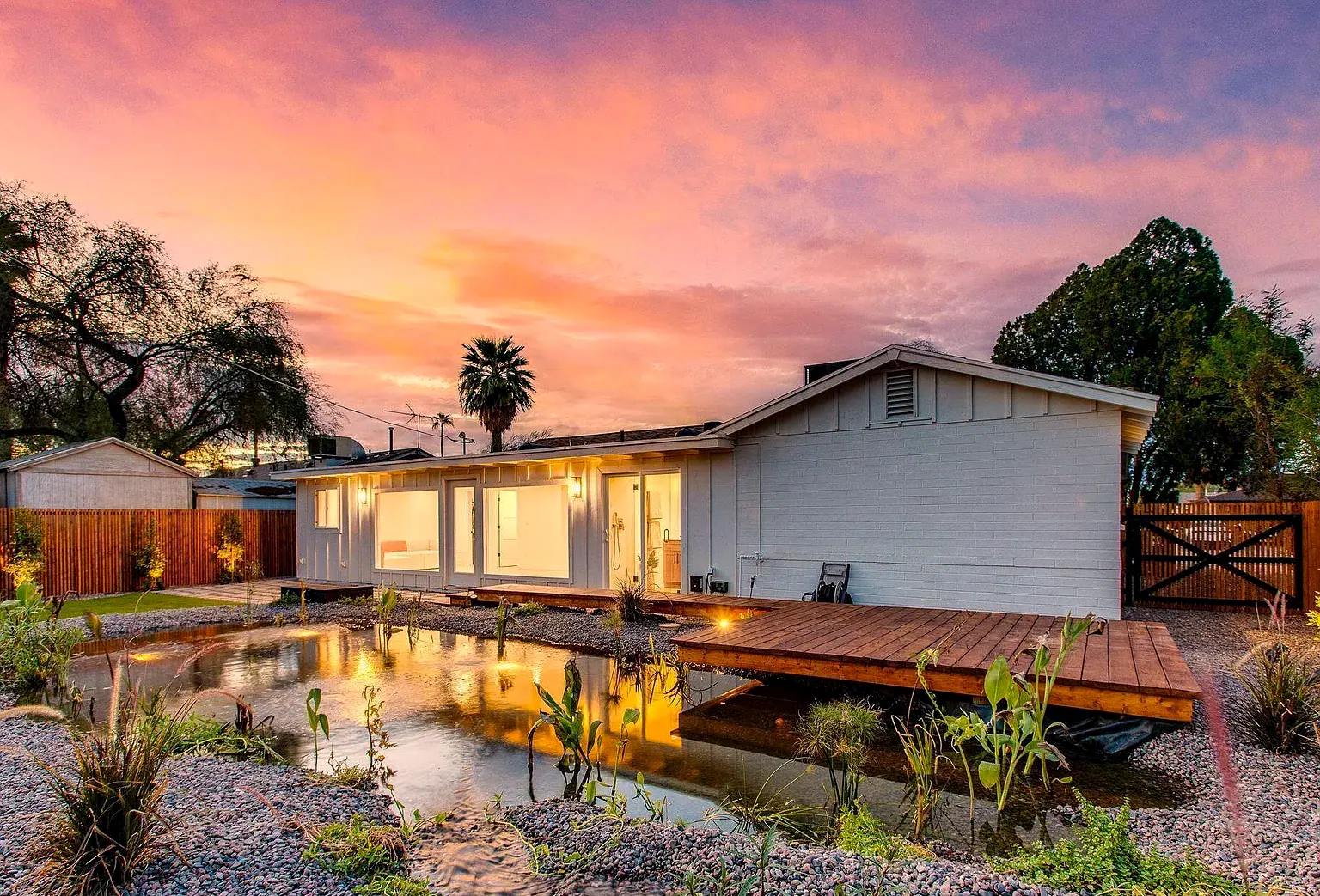 A completed Coveted Contracting renovation at dusk: warm interior light through new sliders, a cedar deck bridging a naturalized pond, pink and orange sky.